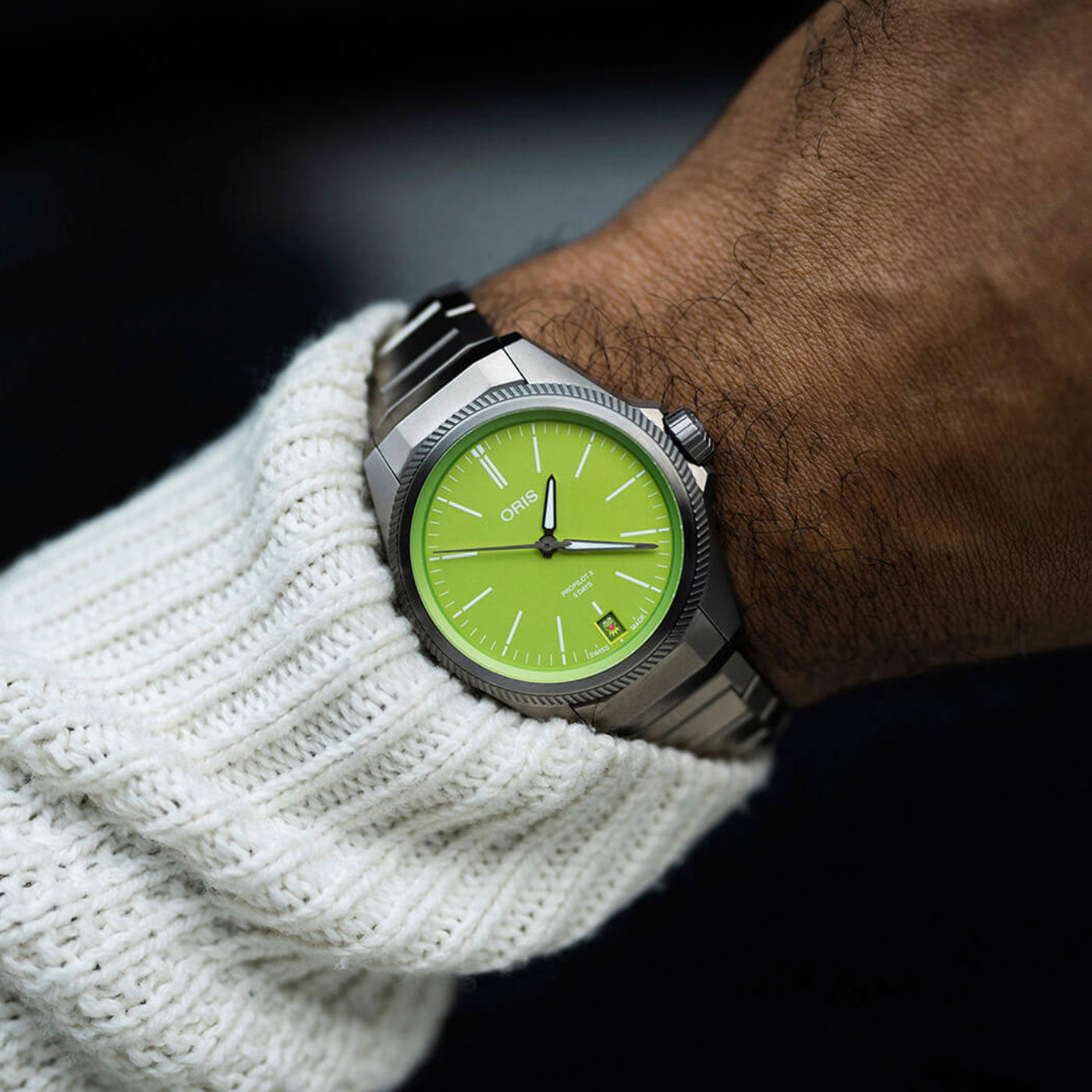 A close-up shows a wrist adorned with an Oris Propilot X Kermit Edition Automatic 39mm Watch, featuring a silver metal band and a bright green dial with white hour markers and ORIS branding. The wearer has on a white knit sweater, set against a dark background.