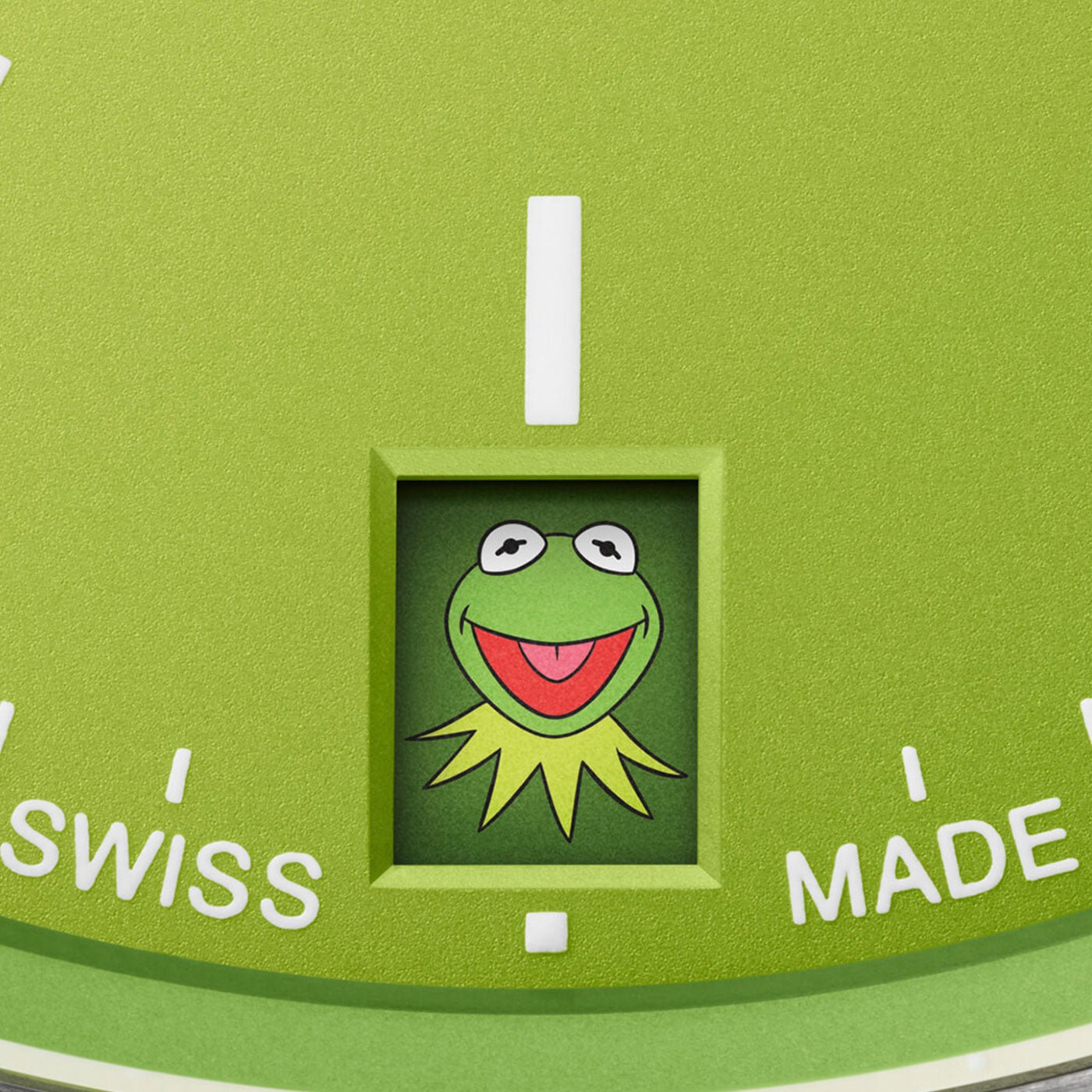 Close-up of an Oris Propilot X Kermit Edition Automatic 39mm watch featuring a green face with a small window showcasing an illustration of a smiling, large-eyed character with a collar. The ORIS brand name and "SWISS MADE" text are partially visible among white markers encircling the face.