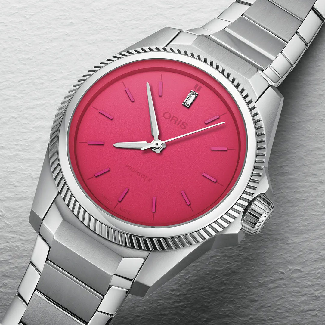 A close-up of the ORIS ProPilot X Miss Piggy Edition Automatic Winding 34mm, featuring a vibrant pink dial, silver hands and hour markers, with a metal bracelet and textured bezel against a light background.