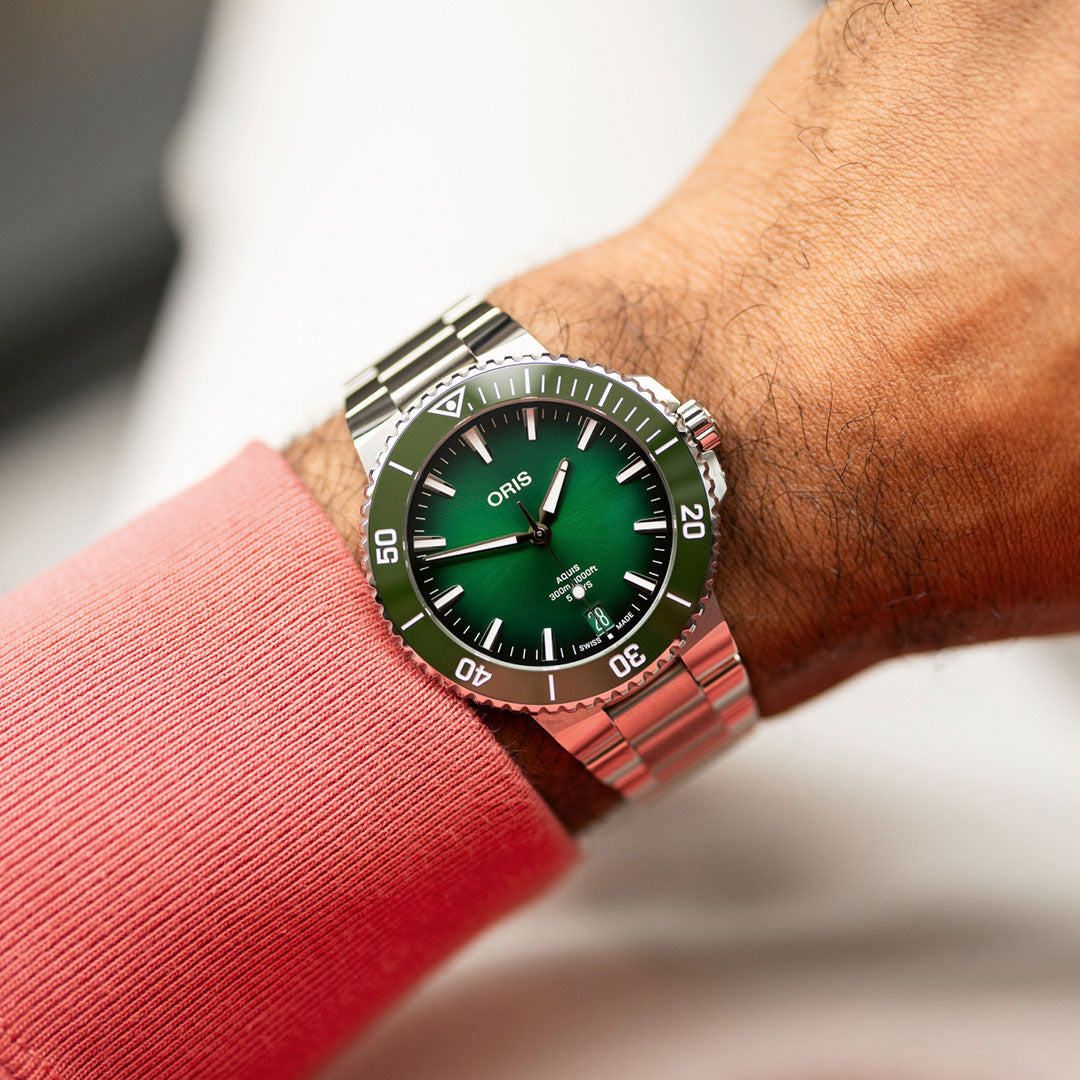 The hand wearing an ORIS Aquis Date Calibre 400 Automatic Winding 43.5mm watch, featuring a sleek green dial and stainless steel bracelet, glints under the light. The pink sleeve complements its innovative design and impressive five-day power reserve.