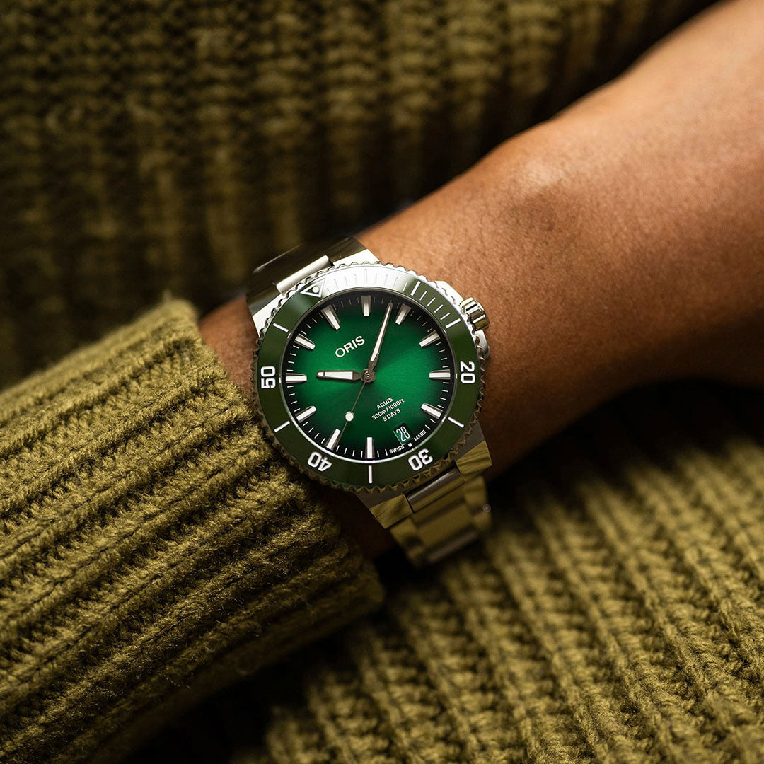 A close-up of a wrist adorned with the ORIS Oris Aquis Date Calibre 400 Automatic Winding 43.5mm Watch in silver showcases its green dial with white markings. The wrist rests gently on a textured olive green sweater, emphasizing the watch's impressive five-day power reserve feature.