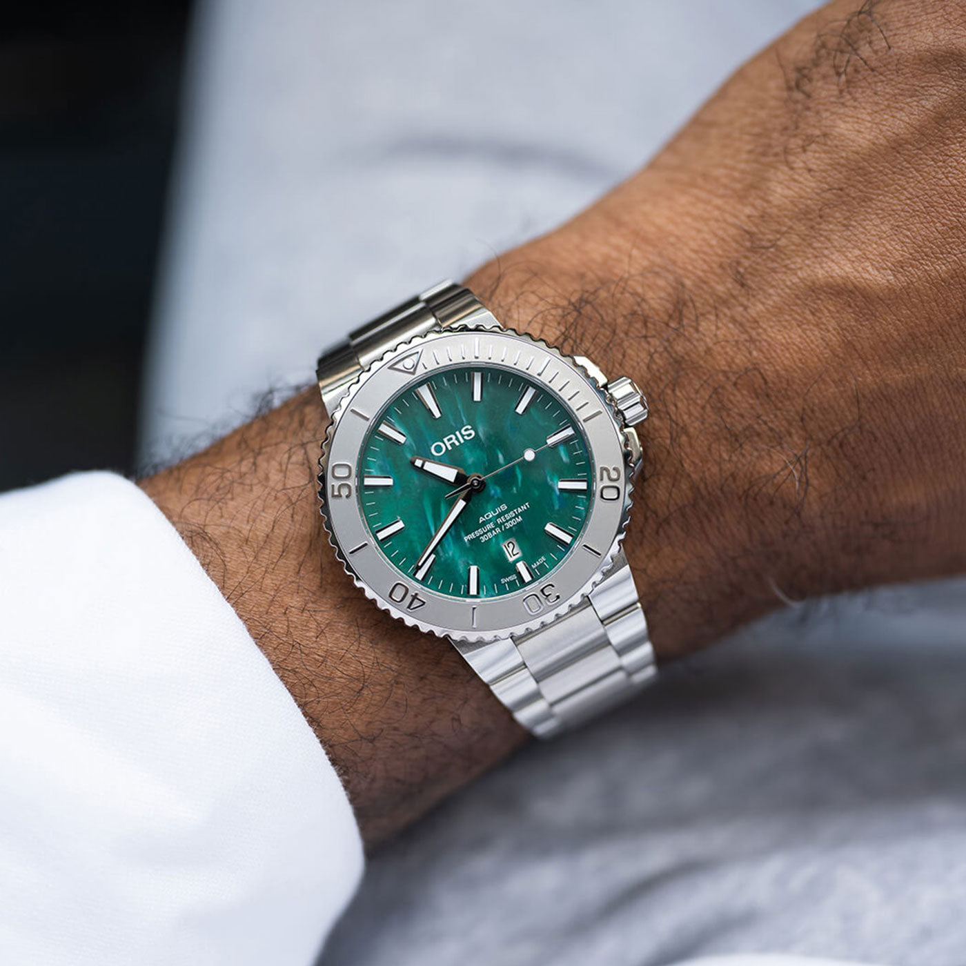 A person's wrist showcases an ORIS Oris Aquis X Bracenet Automatic 43.5mm Watch with a metallic band and a striking green face, standing out against a crisp white shirt. The blurred background emphasizes the watch's exquisite details.