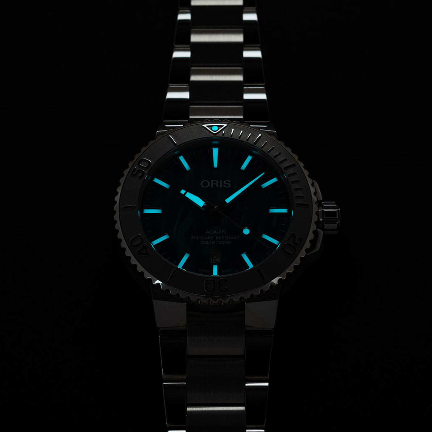 A close-up of the Oris Aquis X Bracenet Automatic 43.5mm watch reveals its luminous blue markers and hands set against a dark background. The watch, with its metal band, prominently features the ORIS name on its dial.