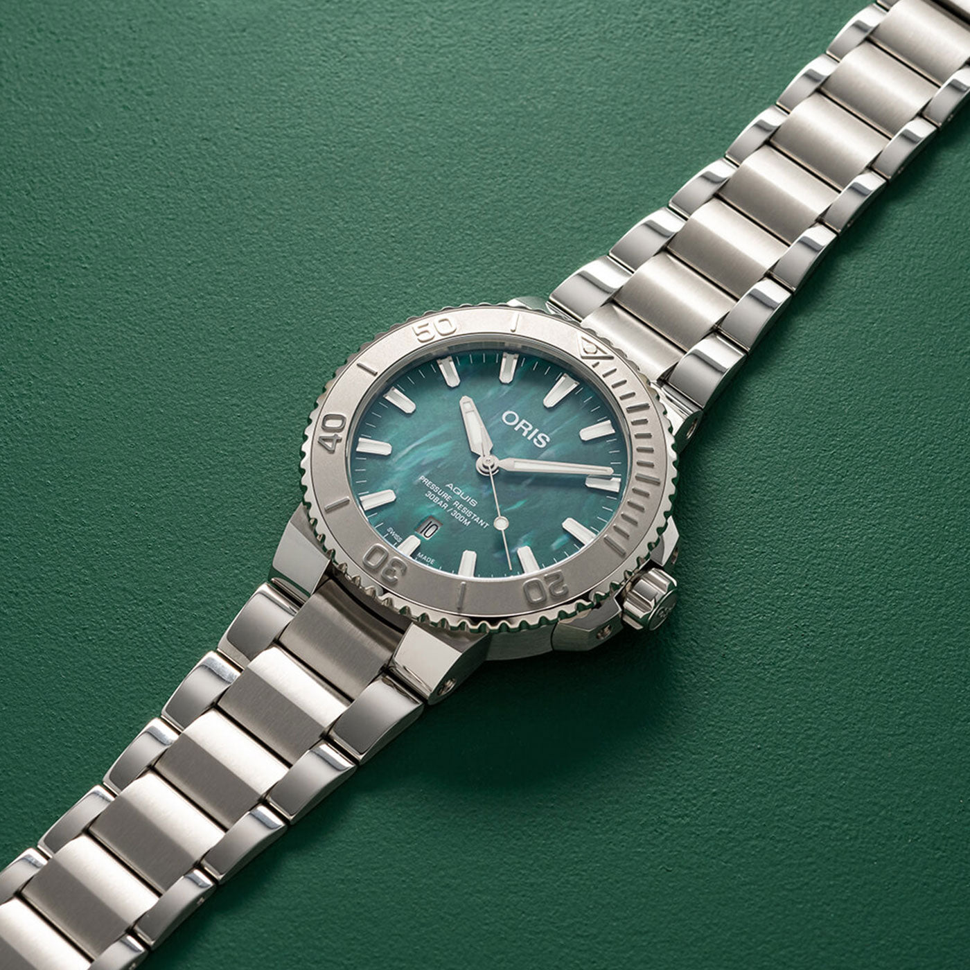 The ORIS Aquis X Bracenet Automatic 43.5mm Watch, featuring a sleek silver metal design with a teal face and white hands, rests diagonally against a dark green backdrop. This sophisticated timepiece showcases a rotating bezel that combines both style and functionality.