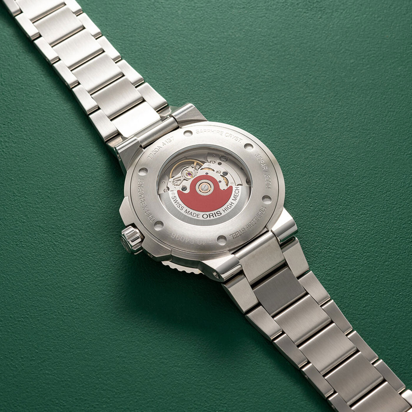The Oris Aquis X Bracenet Automatic 43.5mm Watch, a masterpiece by ORIS, is displayed with its stainless steel back opened to reveal the intricate interior mechanics. Set against a striking green background, the movement stands out with its distinctive red accent. The refined bracelet is also elegantly crafted from stainless steel.