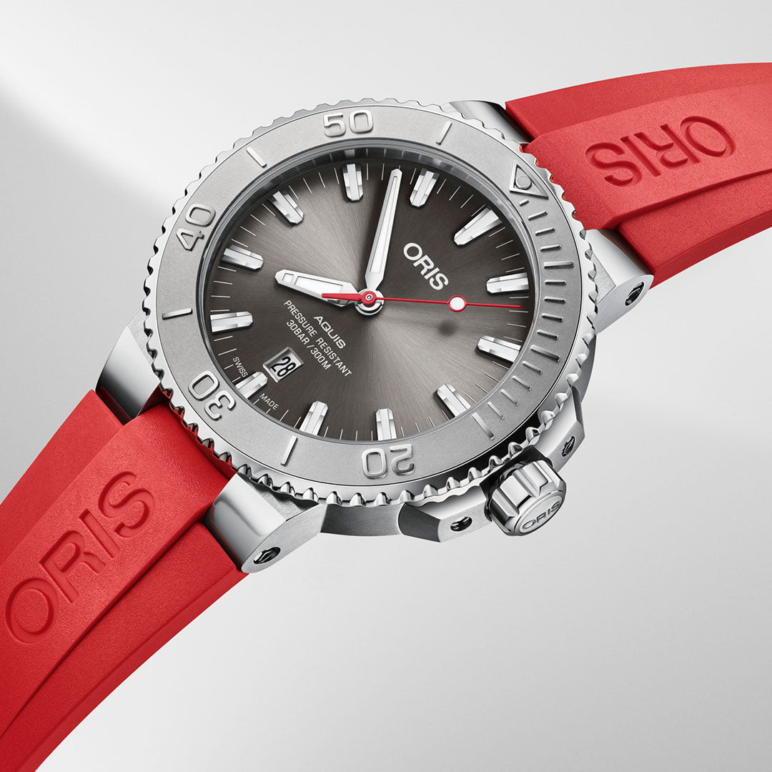 This Oris Aquis Date Relief Automatic Winding 43.5mm Watch, crafted in Switzerland, showcases a sleek gray dial complemented by a silver-tone case. It features a clear date display and luminous markers, and is paired with a vibrant red rubber strap embossed with the "ORIS" logo, adding contrast against its light background.