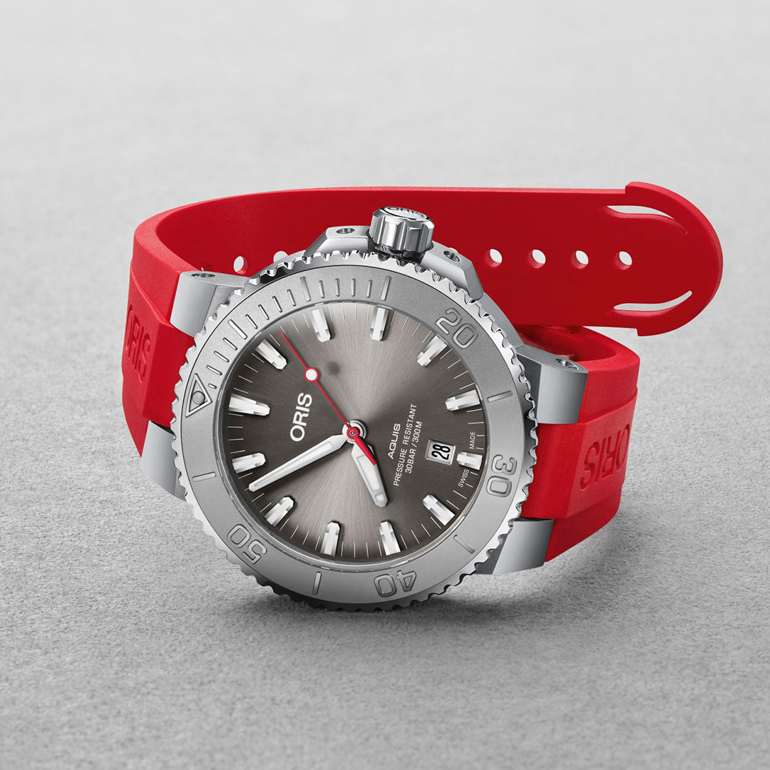 The Oris Aquis Date Relief Automatic Winding 43.5mm Watch by ORIS is a Swiss Made timepiece that showcases a silver dive design with a gray dial and a date display positioned at 3 o'clock. It features water resistance, a robust red rubber strap, and a textured bezel with minute markings, all presented on an elegant light gray surface.