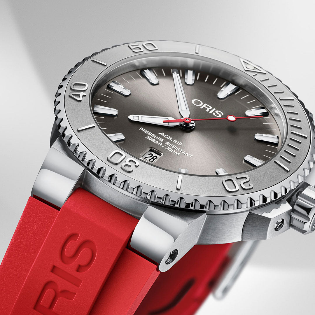 A detailed view of the Oris Aquis Date Relief Automatic Winding 43.5mm watch reveals its red rubber strap, silver bezel with engraved numbers, grey dial featuring luminous hour markers, and a date display. The prominent "ORIS" branding on the dial and strap emphasizes its quality and water resistance.