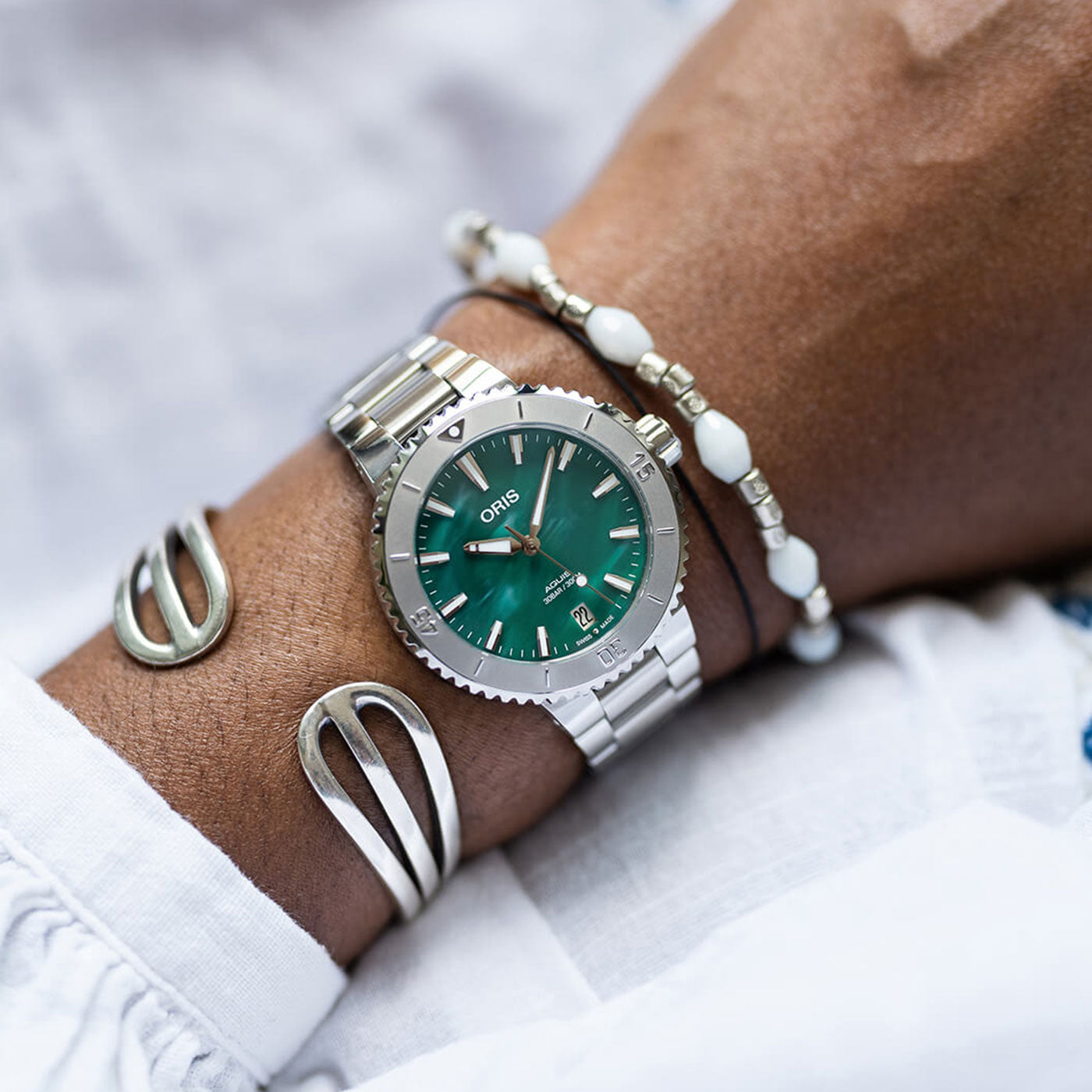 A close-up of a wrist showcases the ORIS Oris Aquis X Bracenet Automatic 36.5mm Watch, featuring a green face and accompanied by two bracelets: one silver with open loops and another adorned with white beads. The stylish ensemble is complemented by a white long-sleeve shirt.