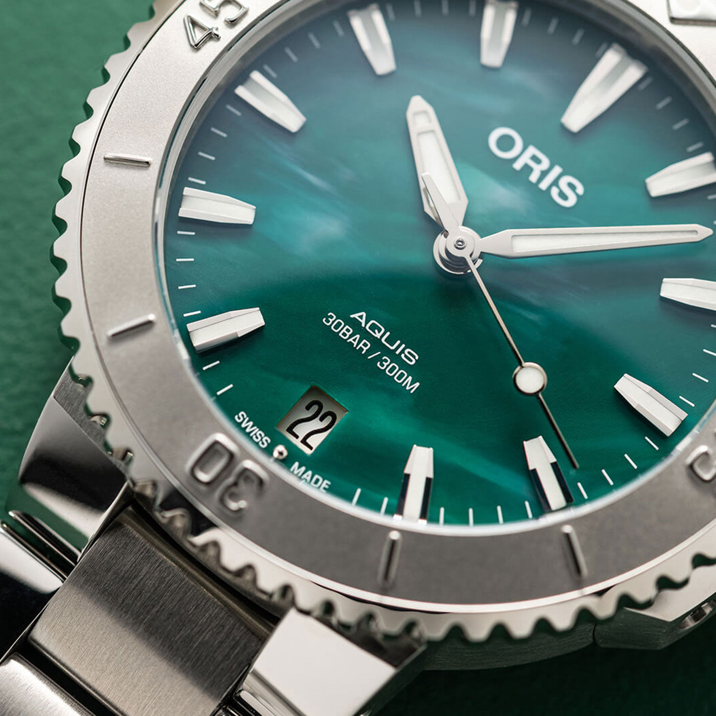 Close-up of a silver Oris Aquis X Bracenet Automatic 36.5mm watch by ORIS, featuring a captivating green dial, white hour markers, and bold hands. The date displays "22," with inscriptions "30BAR/300M" and "Swiss Made." Resting on a green textured surface, this timepiece exemplifies its precise craftsmanship.