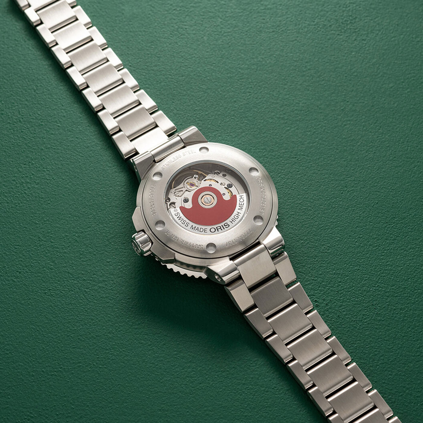 The Oris Aquis X Bracenet Automatic 36.5mm Watch by ORIS features a transparent case back that reveals its intricate mechanical movement. Displayed prominently across a textured green surface, the fully extended metal bracelet highlights the watch's sophisticated craftsmanship.