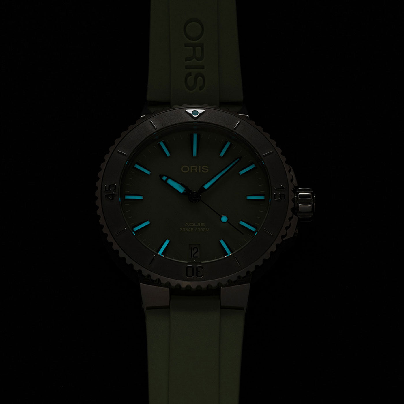 The Oris Aquis Date Automatic Winding 36.5mm Watch features a dark theme with a striking green strap, complemented by luminous blue markers and hands on the dial. The ORIS brand name is prominently displayed on both the watch face and strap, set against a black background that accentuates the watch's distinct features.