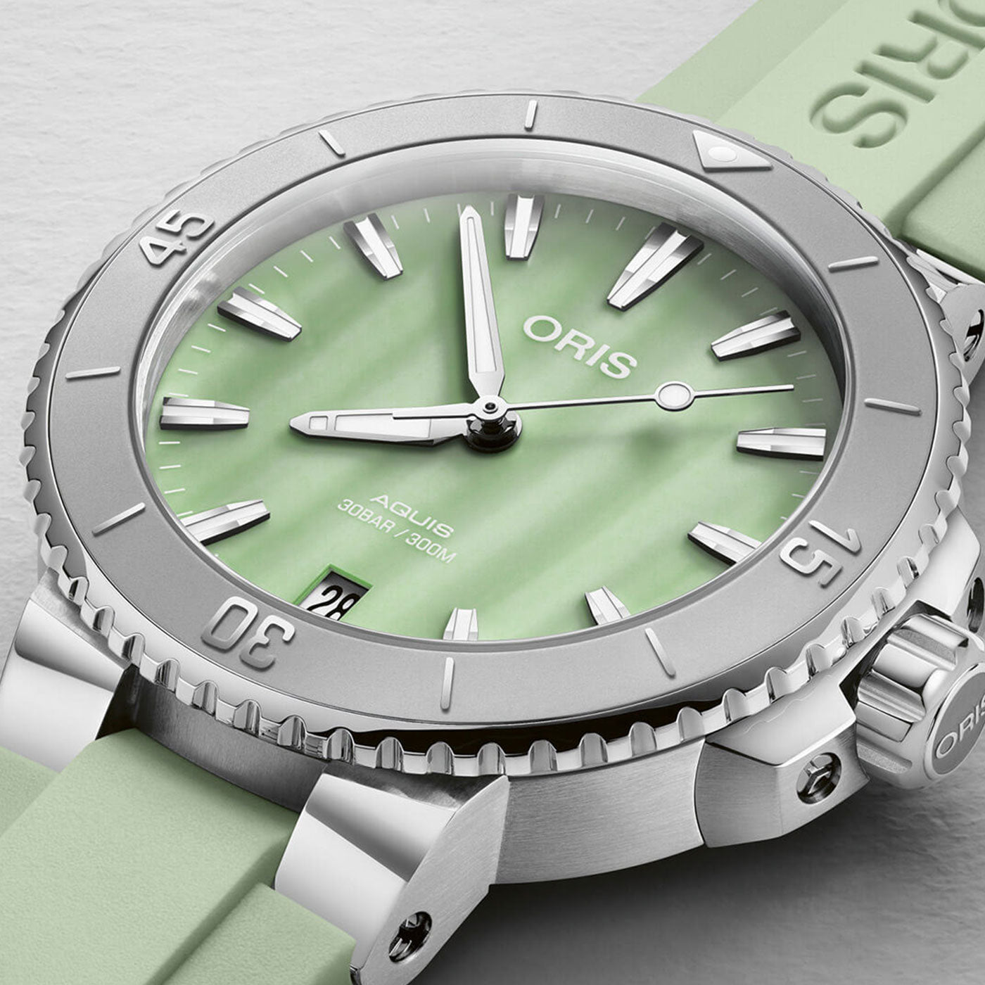 The Oris Aquis Date Automatic Winding 36.5mm Watch by ORIS boasts a silver-toned design with a light green dial and date display at the 6 o'clock position. It includes luminous hands and markers, complemented by a matching light green strap. The bezel numerals enhance its elegant appearance.