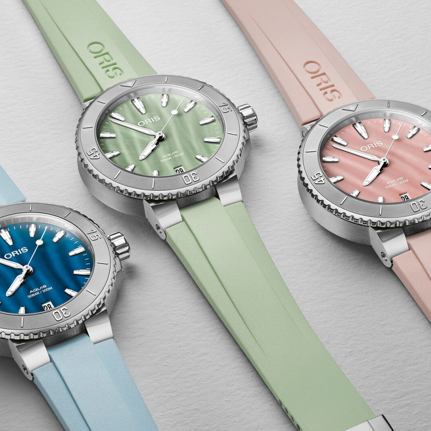A selection of Oris Aquis Date Automatic Winding 36.5mm Watches by ORIS is elegantly displayed on a light surface, highlighting their colorful dials and matching rubber straps in vibrant shades of blue, mint green, and peach. Each watch showcases a silver case with a distinct ridged bezel and bold hour markers that effortlessly capture attention.