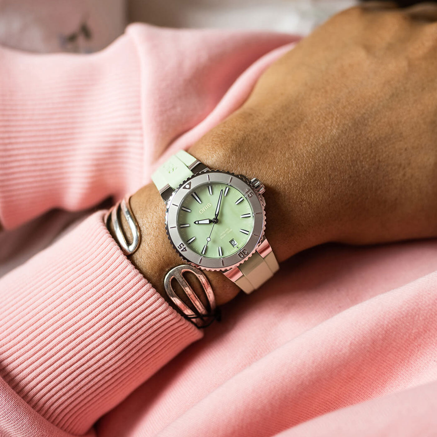 A close-up of a wrist adorned with the Oris Aquis Date Automatic Winding 36.5mm Watch, by ORIS, showcasing a round face with Roman numerals in silver and mint green tones. The person is wearing a pink long-sleeve top, beautifully complemented by two silver bangles.