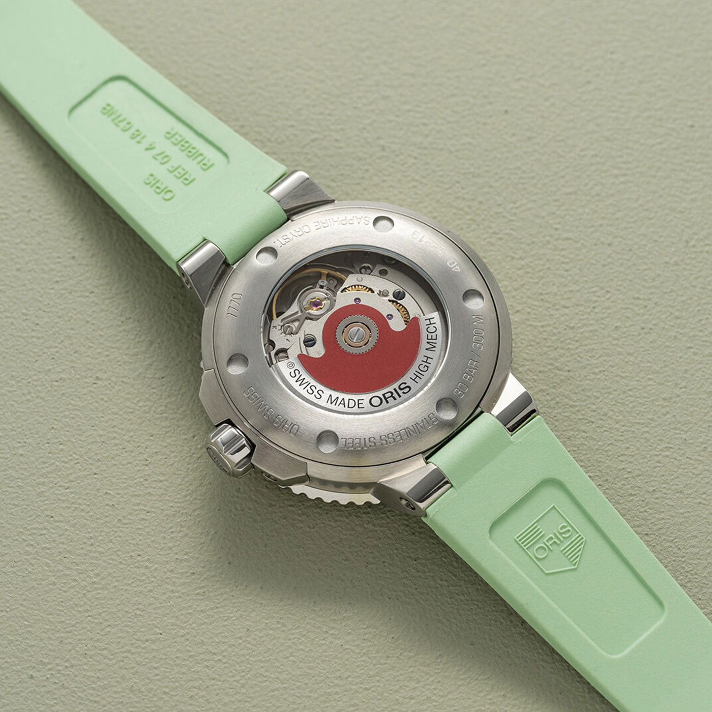 The back of the Oris Aquis Date Automatic Winding 36.5mm Watch reveals a visible mechanical movement with "Swiss Made Oris High Mech." This timepiece is equipped with a light green rubber strap and a polished metal casing, elegantly presented on a matching green surface.