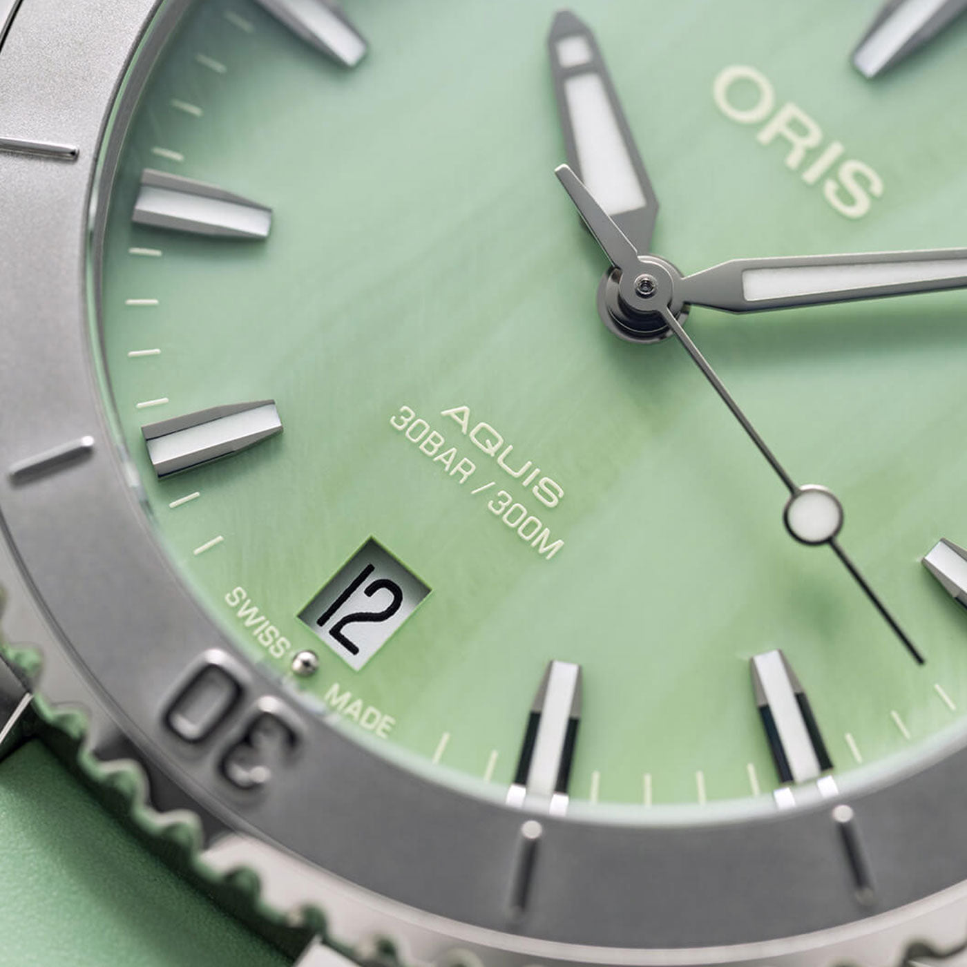 Close-up of the ORIS Aquis Date Automatic Winding 36.5mm watch face, highlighting the brand name, model, and "Swiss Made." It has a date window showing "12" and is water-resistant up to 300m/30bar. The textured dial features silver hands and markers that exude elegance and durability.