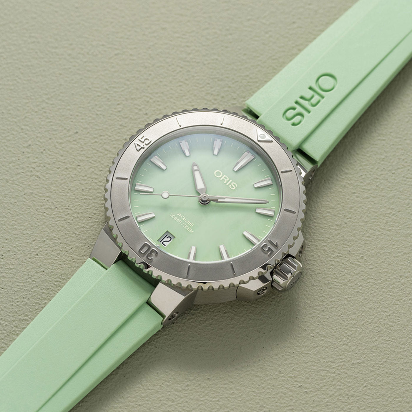 The Oris Aquis Date Automatic Winding 36.5mm Watch by ORIS showcases a stylish silver diving design with a light green face and matching rubber strap, featuring luminous hands and markers. It is set against a textured, vibrant backdrop, perfectly embodying elegance and adventure for ocean enthusiasts.