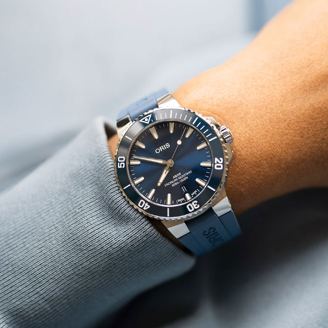 A close-up of a wrist wearing the ORIS Aquis Date Automatic Winding 41.5mm Watch, featuring a blue rubber strap, silver case, white markers, and rotating bezel against a light blue sleeve—a refined luxury toolwatch.