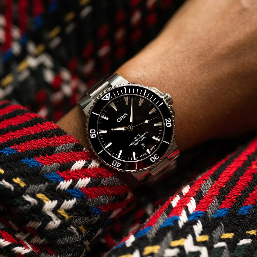 A close-up shot captures a wrist elegantly adorned with an ORIS Oris Aquis Date Automatic Winding 43.5mm Watch, featuring a silver finish, black face, and white indices. The wearer stylishly complements it with a vibrant sweater that boasts red, black, white, and blue zigzag patterns.