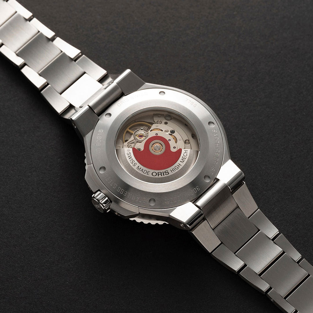 The ORIS Aquis Date Automatic Winding 43.5mm Watch features a stainless steel sports design with a transparent case back that showcases intricate gears and a red rotor marked "Swiss Made Oris High Mech." Its ceramic insert adds elegance, making it stand out beautifully on a dark surface.