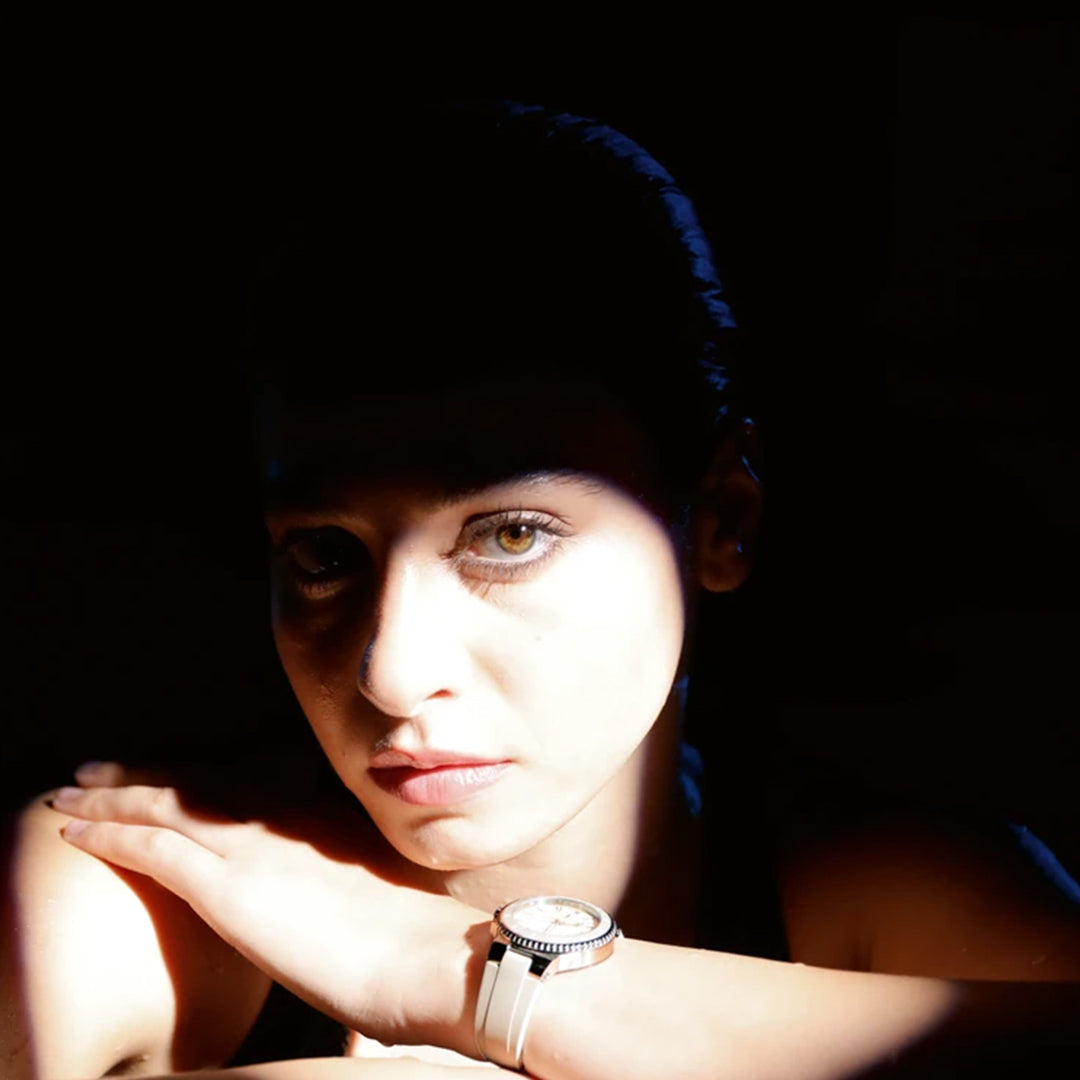 A person, lit by bright light with contrasting shadows on their face, rests their chin on their hands. They're wearing an ORIS Aquis Date Automatic Winding 36.5mm watch and looking directly at the camera against a dark background, enhancing the light's effect.