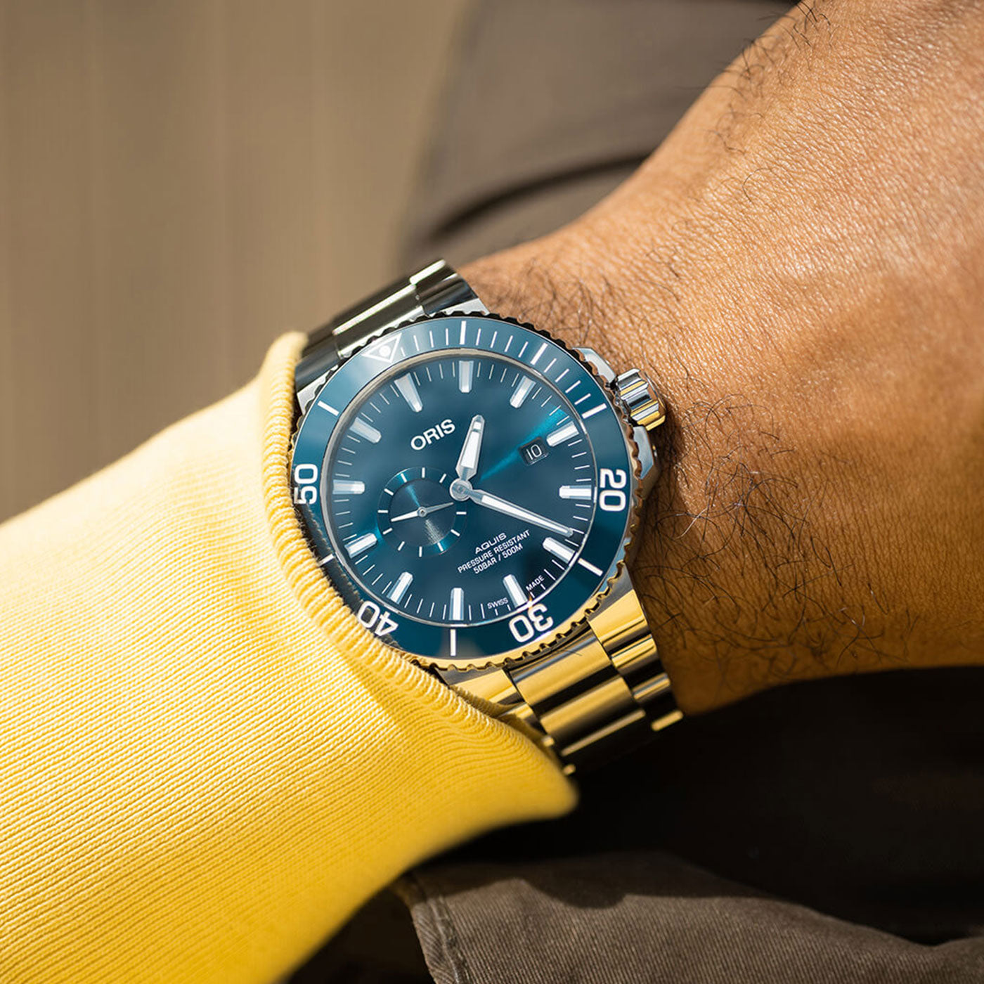 A person is wearing the Oris Aquis Small Second Date Automatic 45.5mm Watch, featuring a stainless steel band and a deep blue dial with minute markings and two subdials. The hand rests on a yellow sleeve, displaying the brand name "ORIS" against a blurred background.