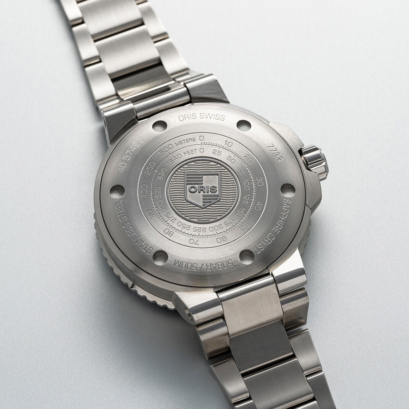 Close-up of a silver metal wristwatch, emphasizing the back of the case. The case displays engraved details including "ORIS" and water resistance information up to 300 meters. The Oris Aquis Small Second Date Automatic 45.5mm Watch features a polished metal bracelet.