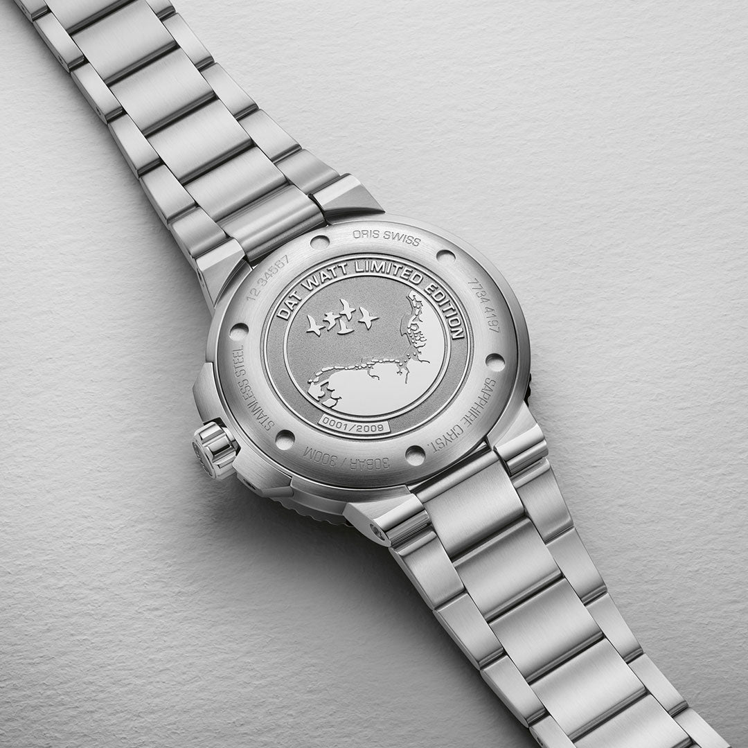 The image features the reverse side of the ORIS Aquis Dat Watt Limited Edition II Automatic Winding 43.5mm Watch, displaying a detailed engraved metal case back. The elaborate engraving includes a world map and three birds representing conservation efforts, along with the inscription: "ORIS DAT WATT LIMITED EDITION II," marked as "0201/2009." This remarkable watch is paired with a sophisticated metal link band.