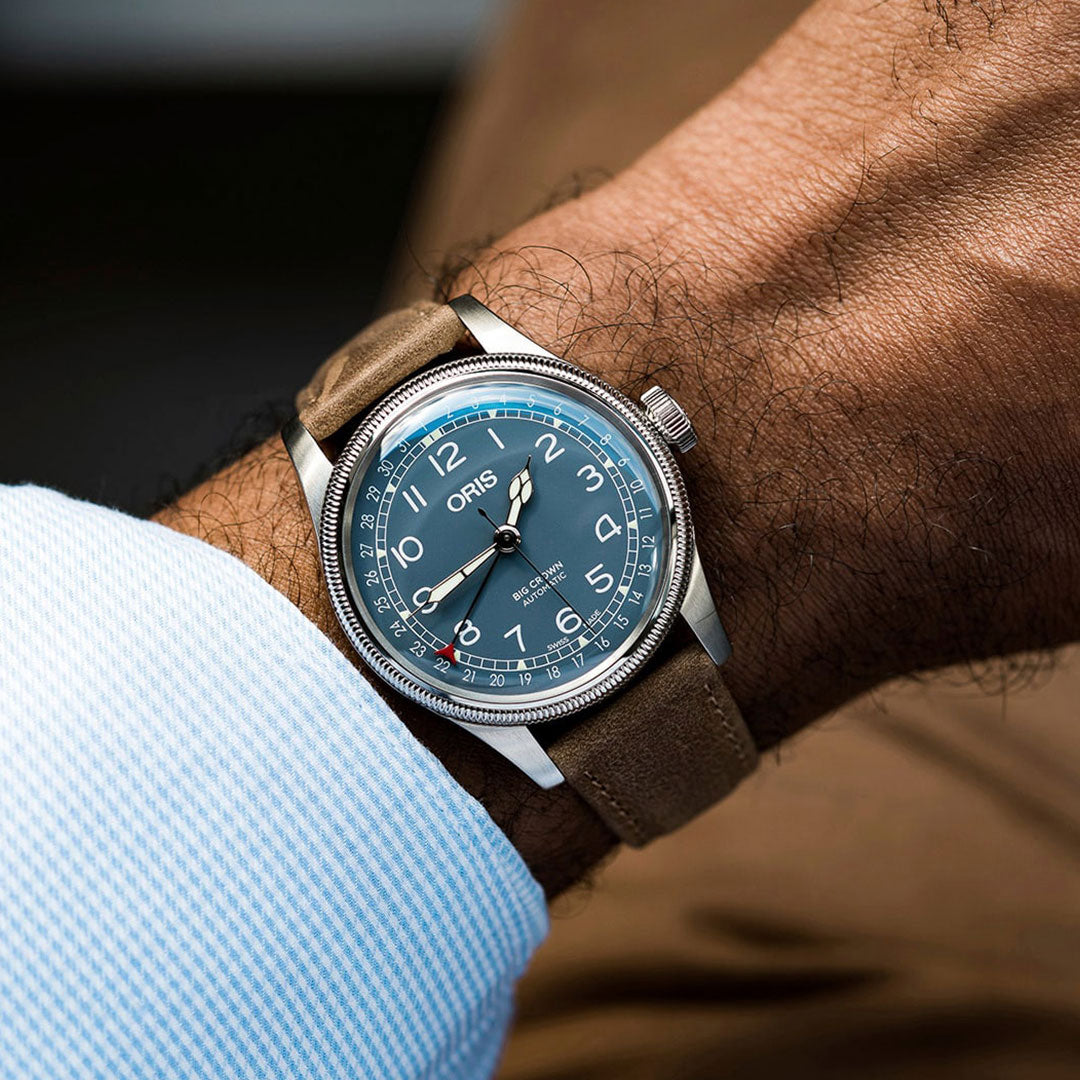 A close-up of a wrist adorned with the Oris Big Crown Pointer Date Automatic Winding 44mm Watch by ORIS, showcasing its brown leather strap and blue face with white numerals and date indicator, complemented by an oversized crown. The individual is wearing a light blue textured shirt that harmonizes perfectly with the watch's sophisticated style.