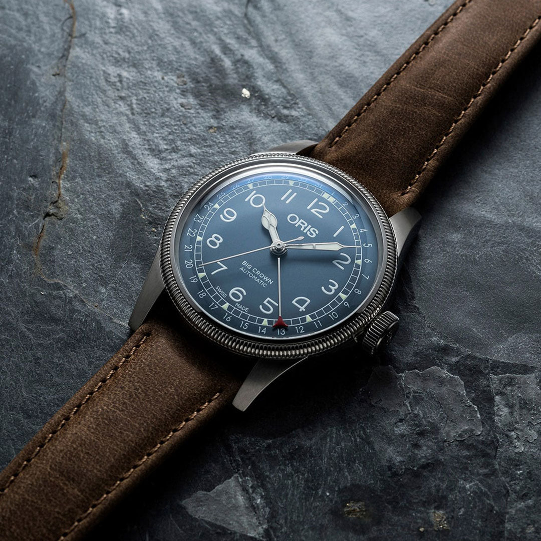 An Oris Big Crown Pointer Date Automatic Winding 44mm Watch for men with a brown leather strap and a blue dial displaying white numerals. The brand name "ORIS" is prominently featured on the watch face, with its oversized crown adding to its rugged charm as it rests elegantly on a textured dark surface.