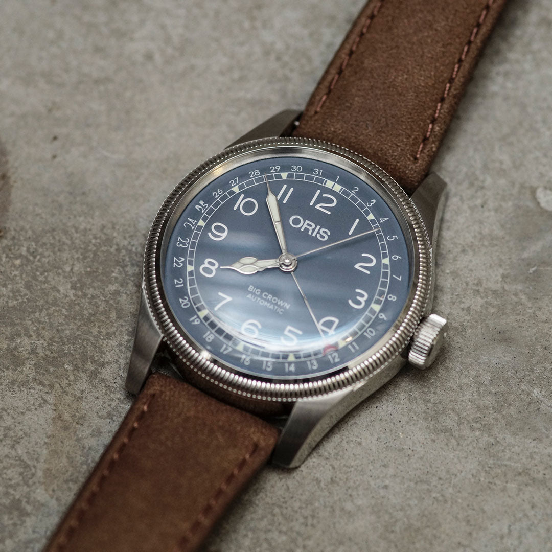A watch from ORIS, the Big Crown Pointer Date Automatic Winding 44mm, with a silver case and brown leather strap, rests on a gray stone surface. Its dark face showcases white numerals and hands, while prominently displaying the brand "ORIS" along with "Big Crown Pointer Date" and "Automatic." This pilot's watch also features an oversized crown for convenient adjustments.