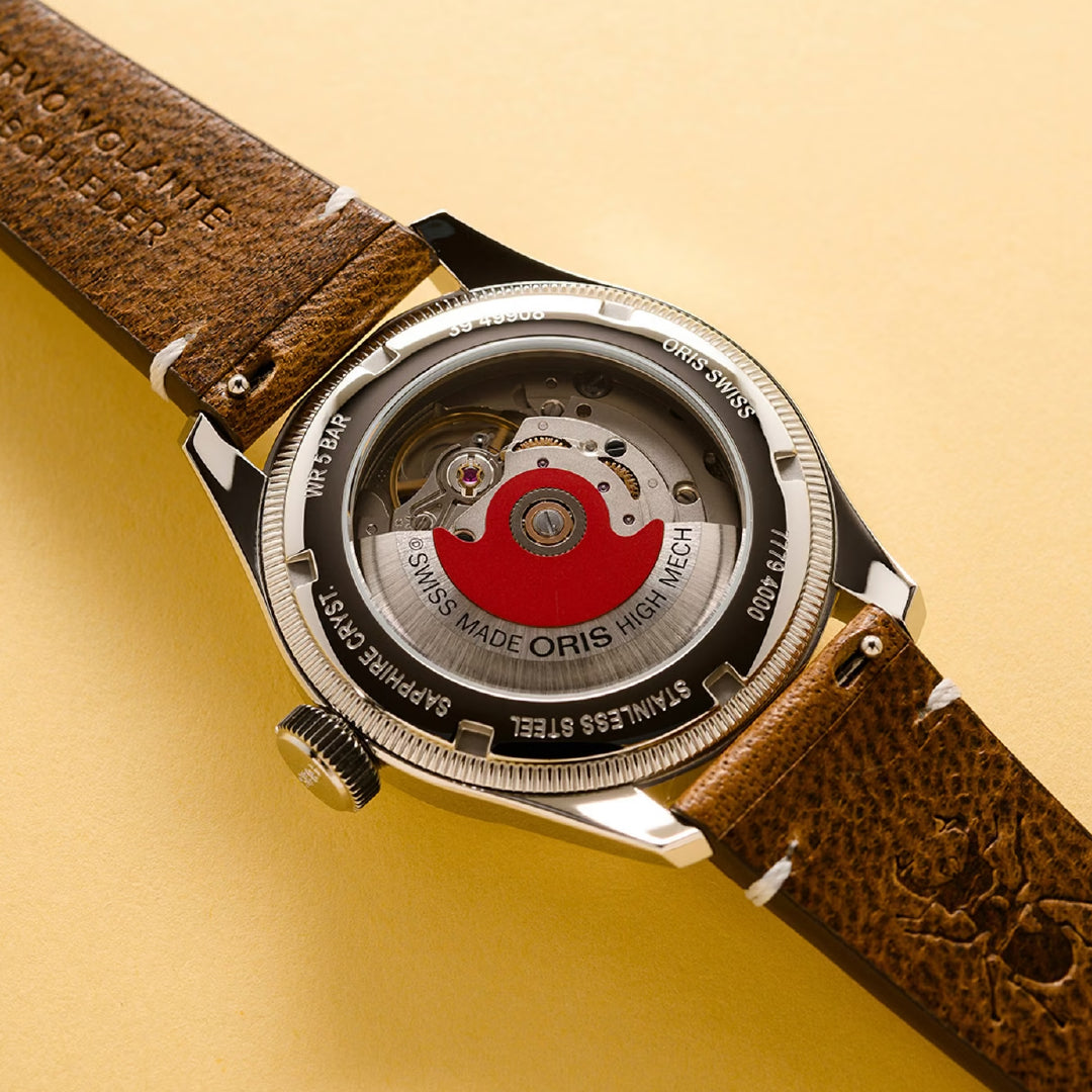 The Oris Big Crown X Cervo Volante 38mm Watch has a transparent back case revealing a red rotor and "Swiss Made" etching. It boasts an eco-friendly deer leather strap, offering an elegant design against a light yellow background.