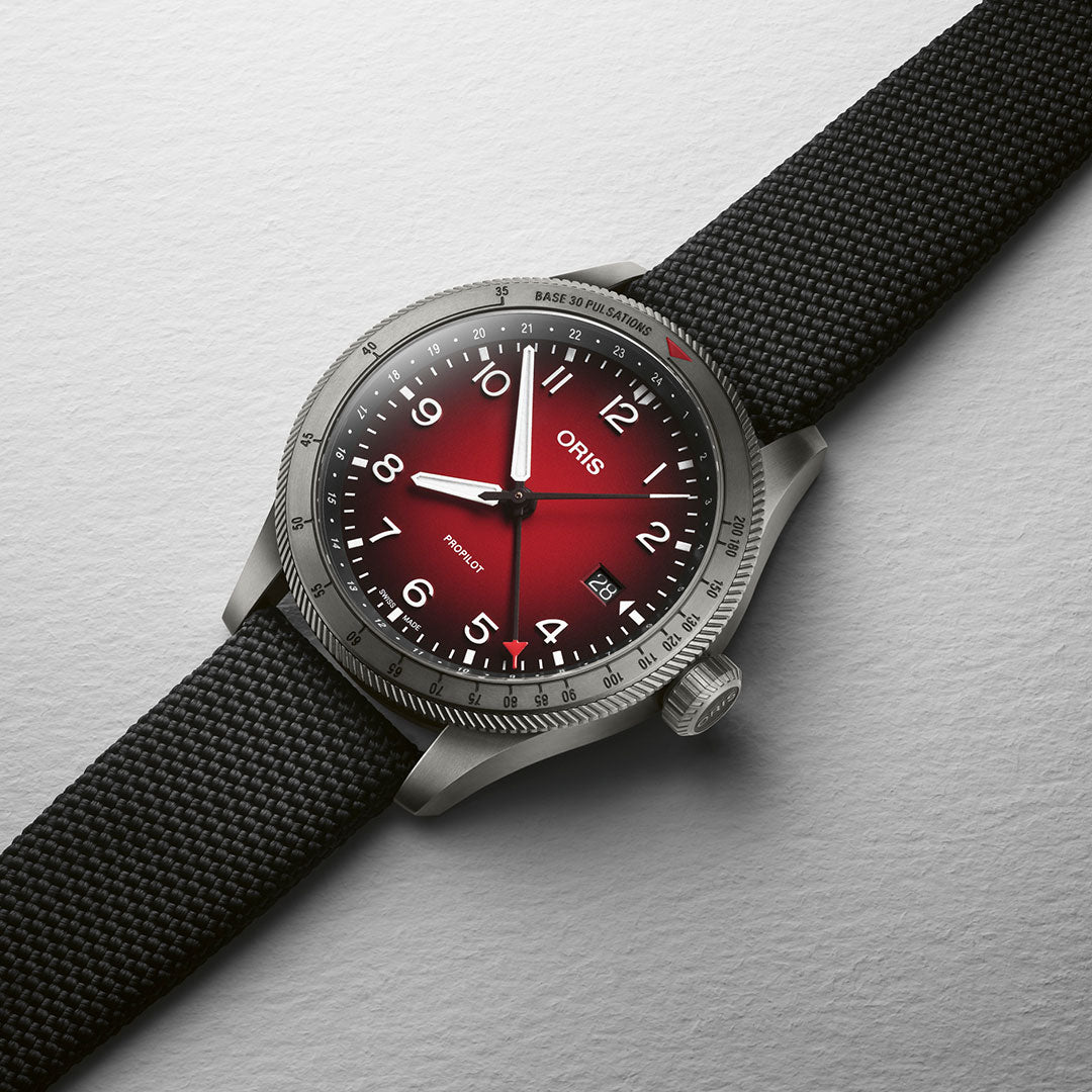 The Oris Propilot GMT Automatic 41.5mm Watch by ORIS showcases a metallic case paired with a striking red gradient dial, highlighted by white numerals and hands, and includes a date display positioned at four o'clock. Its silver bezel is well-matched with a black fabric strap, making it an excellent choice for adventurers requiring accurate second timezone tracking.