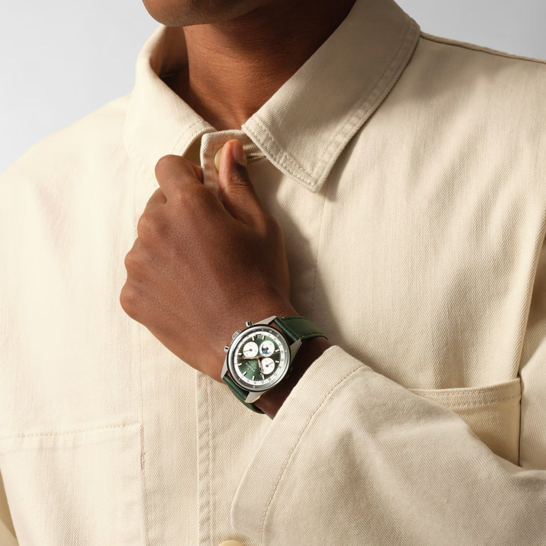A person in a beige jacket adjusts their collar, revealing a ZENITH Chronomaster Original El Primero 3610 on their left wrist. The green watch with a silver bezel and high-frequency chronograph features two subdials against a neutral, minimalist backdrop.