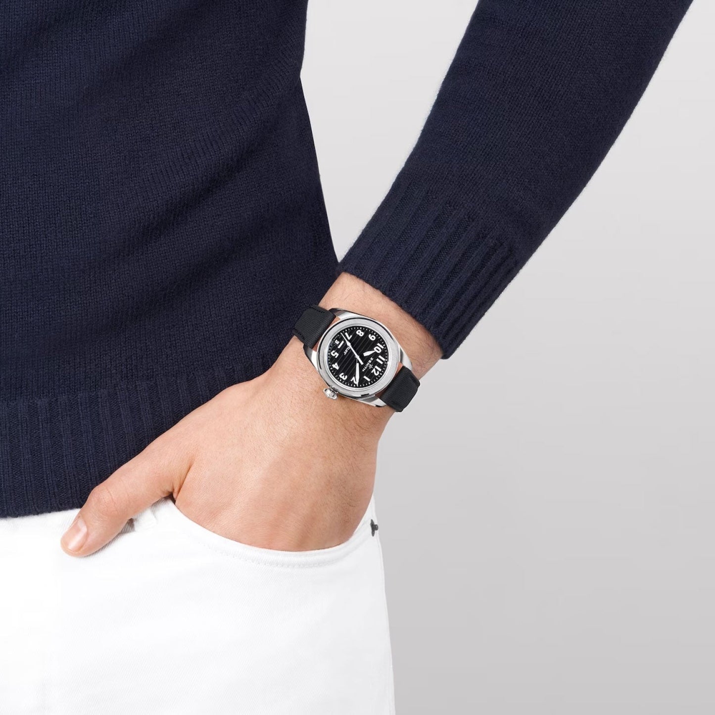 A person in a dark sweater and white pants is donning the ZENITH Pilot El Primero 3620 Automatic 40mm Watch, featuring a round face with a black ceramic case and white numbers. Their hand rests in their pocket against a light gray background.