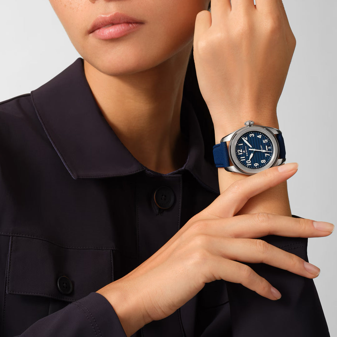 A person in a dark jacket showcases the ZENITH Pilot Automatic El Primero 3620 watch, featuring a large round face with a dark blue strap, white hands and numerals. The oversized crown is subtly highlighted as they gently touch their wrist against a neutral background.