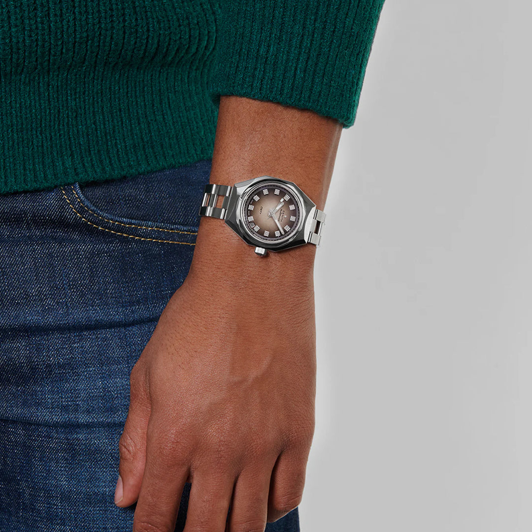 The person is casually dressed in a green knit sweater and blue jeans, showcasing a ZENITH Defy Revival Elite 670 37mm Watch with a round, dark face and metal band. Their relaxed arm against a neutral backdrop beautifully highlights this limited edition Zenith piece.