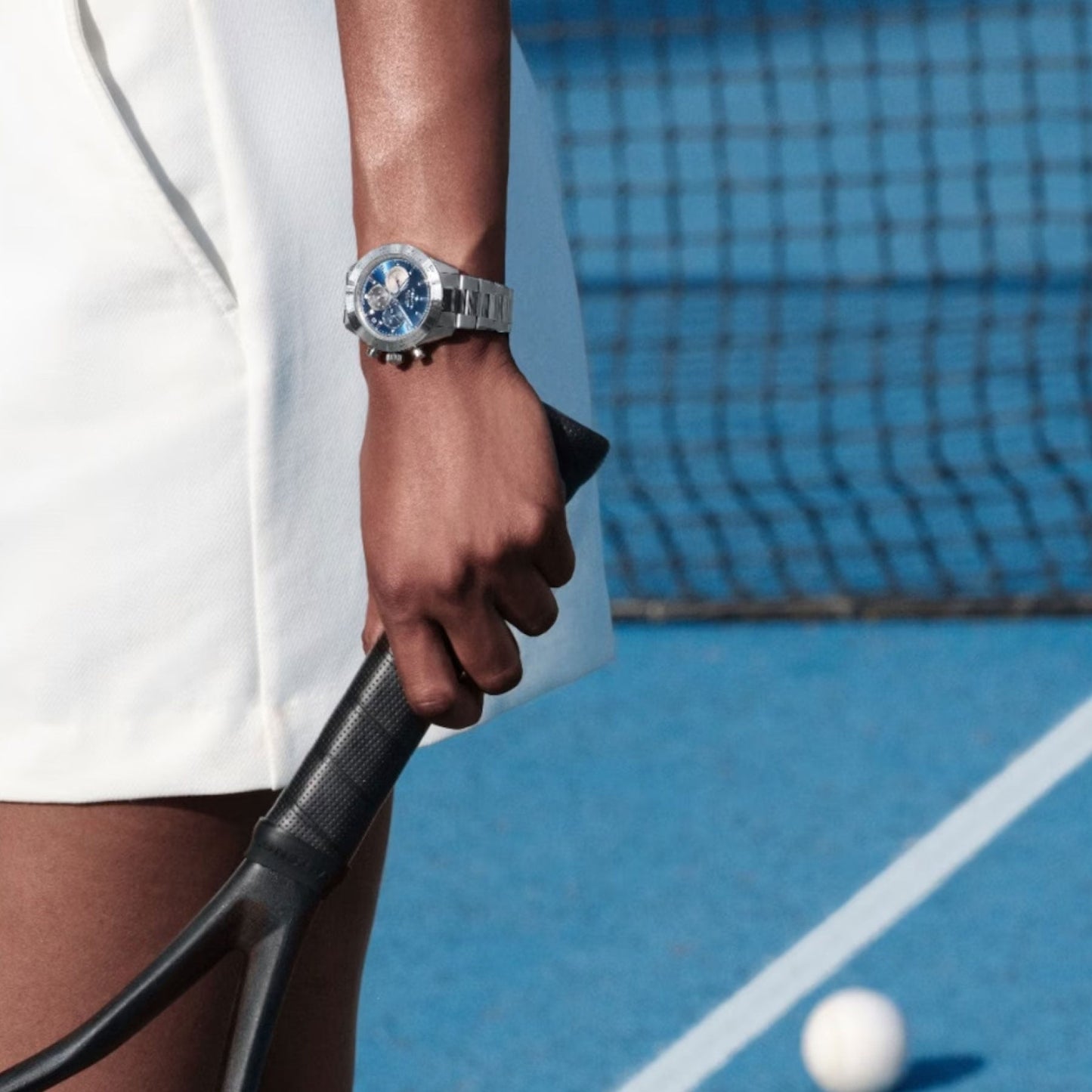 A person dressed in white sports attire is holding a tennis racket on a blue tennis court, with a ball nearby. Adorning their wrist is the Zenith Chronomaster Sport El Primero 3600 Automatic Chronograph 41mm Watch.