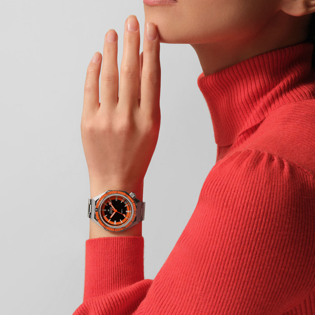 A person wearing a bright orange turtleneck is resting a hand against their face, showcasing the stylish ZENITH Defy Revival Shadow Elite 670 Automatic 37mm Watch, featuring an orange and black dial on a metallic strap. The background is plain and light gray.