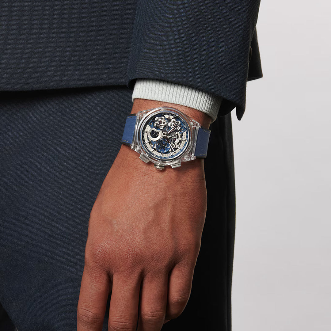 A person wearing a dark suit showcases the Zenith ZENITH Defy Double Tourbillon El Primero 9020 46mm Watch, featuring an intricate, transparent dial and blue strap. The sapphire case reveals internal gears, highlighting its detailed tourbillon craftsmanship.