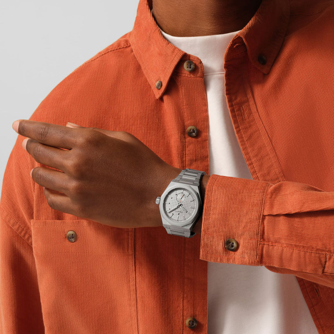 A person in an orange button-up and white T-shirt gestures with their left hand, displaying the Zenith DEFY Skyline C.X. El Primero 3620 SK 41mm Watch—Limited Edition of 200 pieces—known for its precision timekeeping and sleek silver design.