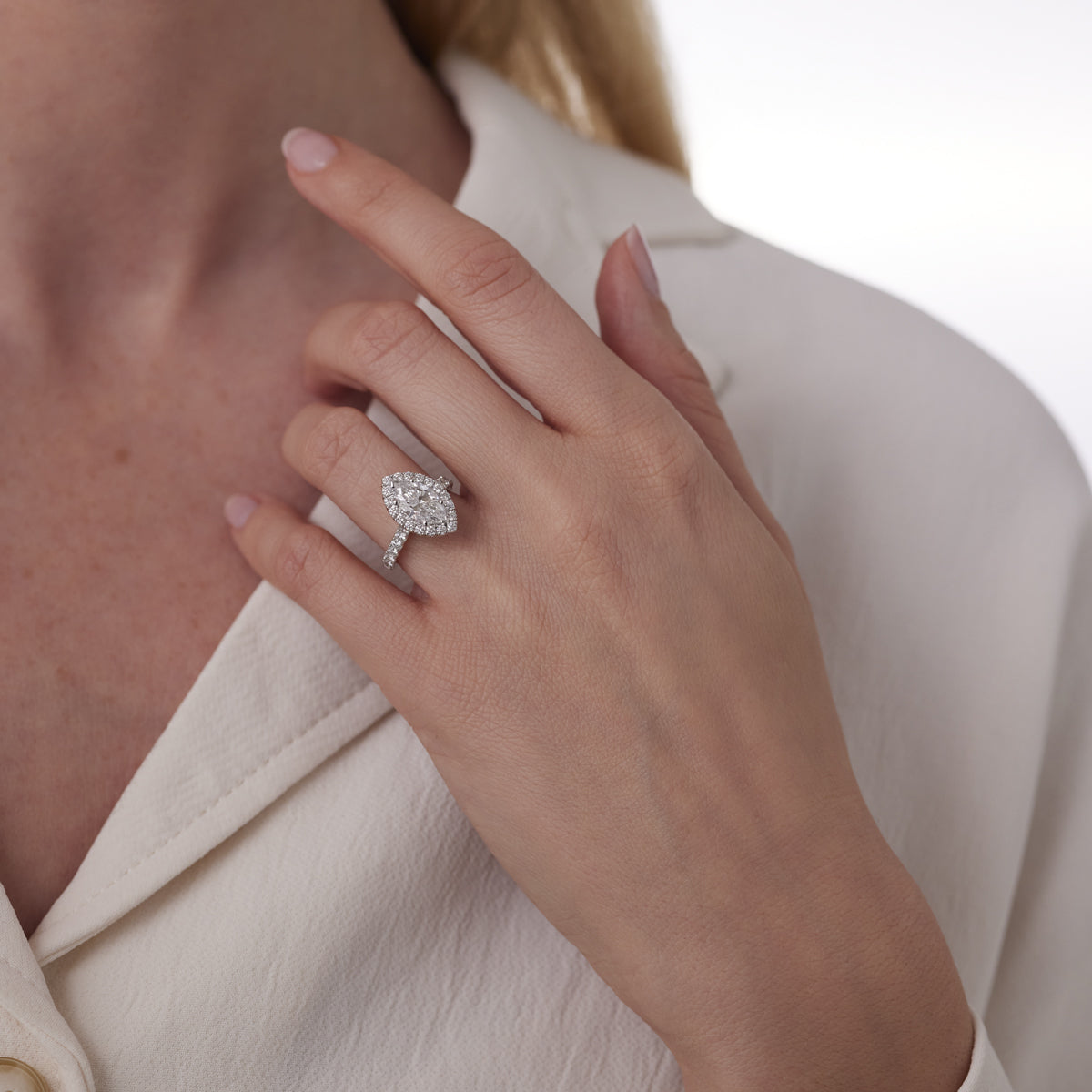 A woman’s hand elegantly presents a Raffi&Co.® 14K White Gold Marquise Halo Diamond Engagement Ring. This exquisite piece, featuring a prominent marquise-cut stone encircled by smaller gems, shines brilliantly against white fabric, radiating elegance and sophistication.