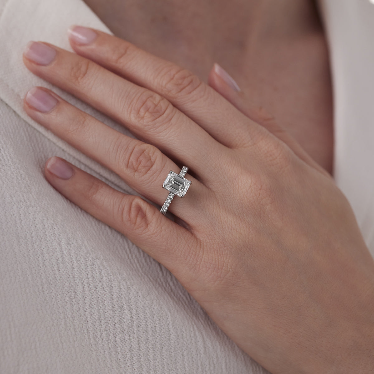 A close-up shot features a hand adorned with the Raffi&Co® 14K White Gold Emerald Hidden Halo Diamond Engagement Ring. The textured band accentuates the GIA-certified gem, while the hand gently rests against a light fabric, highlighting well-manicured nails with a natural polish.