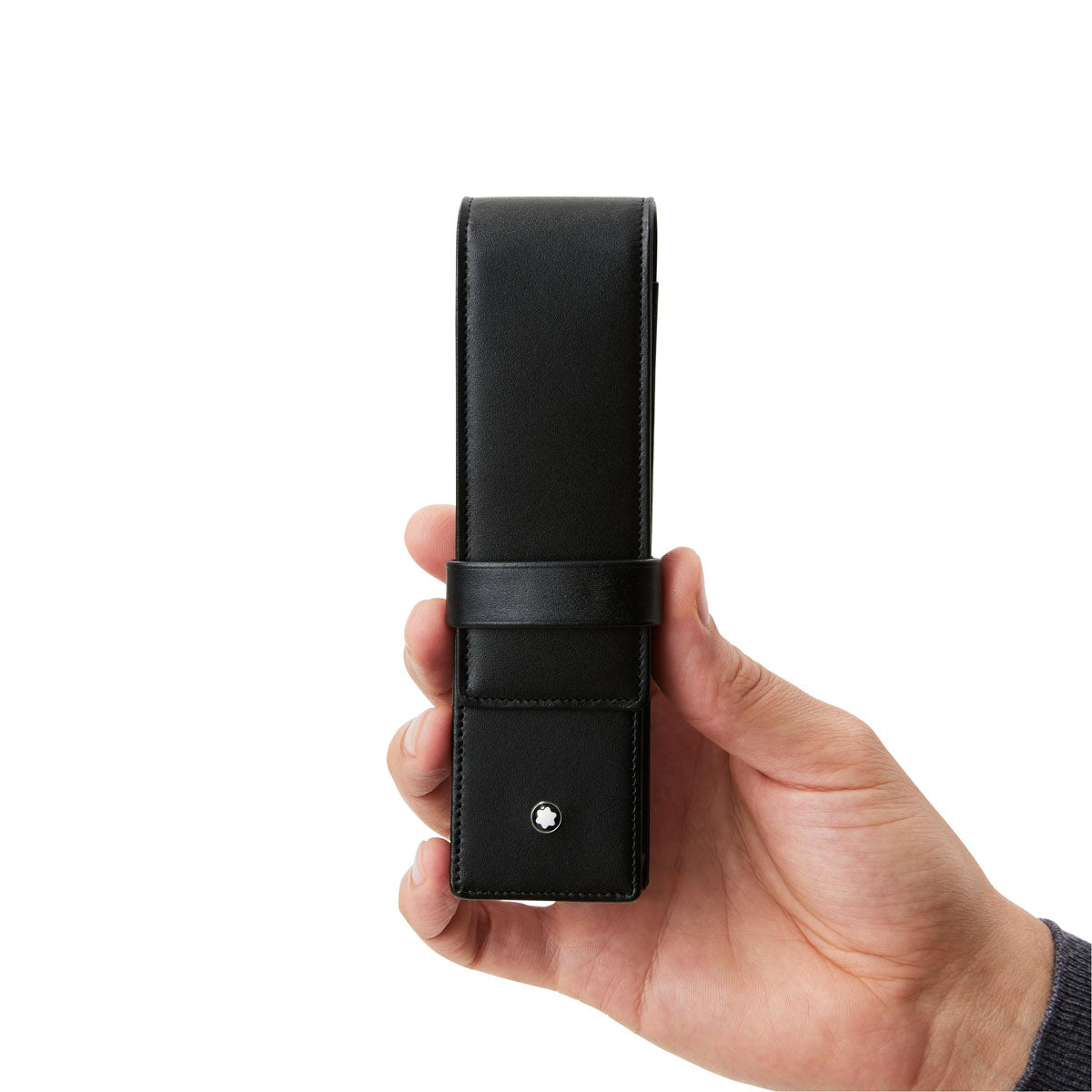 A hand holds a Montblanc Meisterstück Black Leather Pen Pouch, showcasing its sleek design and small button detail near the bottom. The pouch features a strap for closure and displays a minimalist aesthetic against a plain white background.
