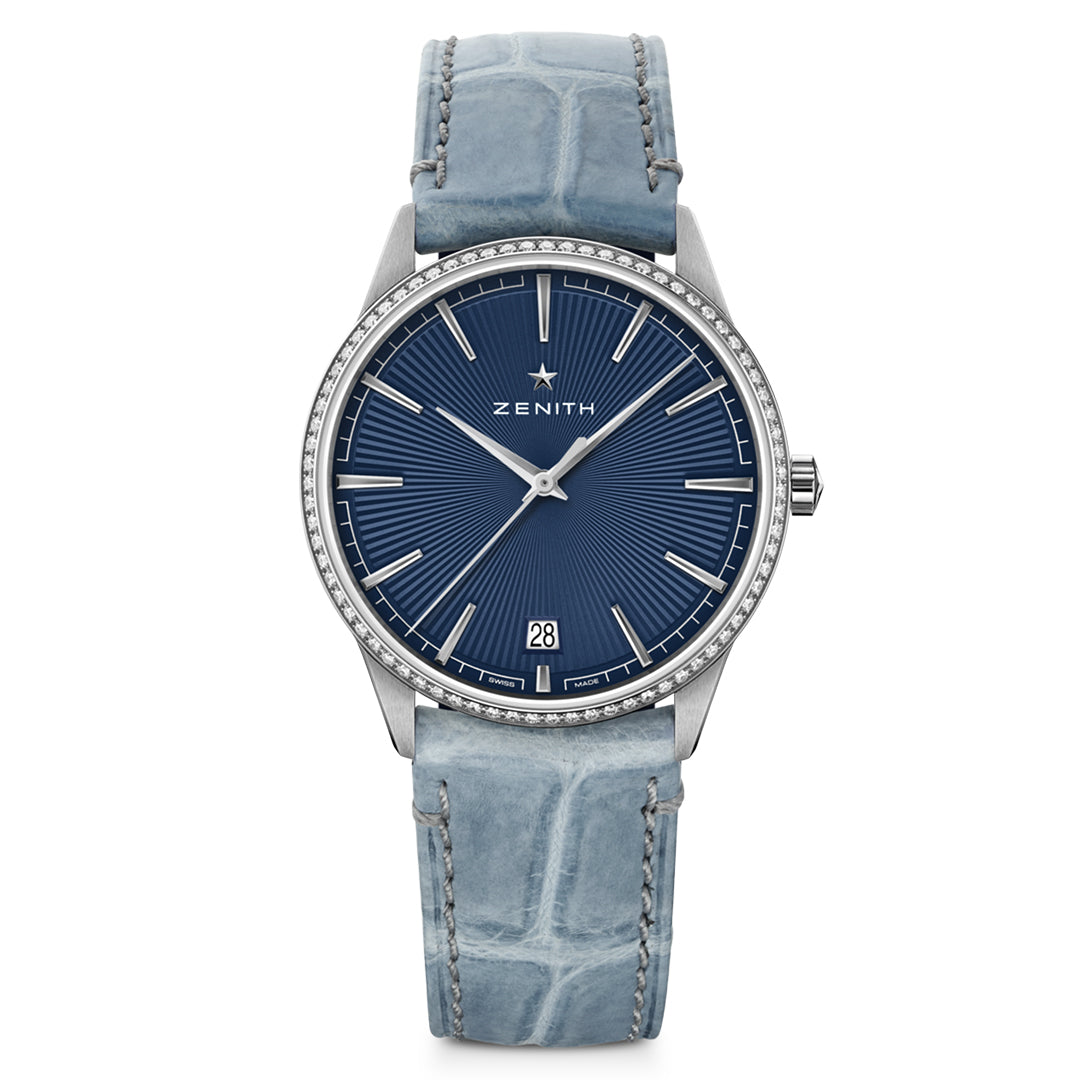 The ZENITH Elite Classic Elite 670 36mm watch features a luxury design with a blue dial, silver hour markers, and brand logo below the 12 o'clock position. It has an automatic winding mechanism, date display at 6 o'clock, a silver crystal-adorned bezel, and a textured light blue leather strap.