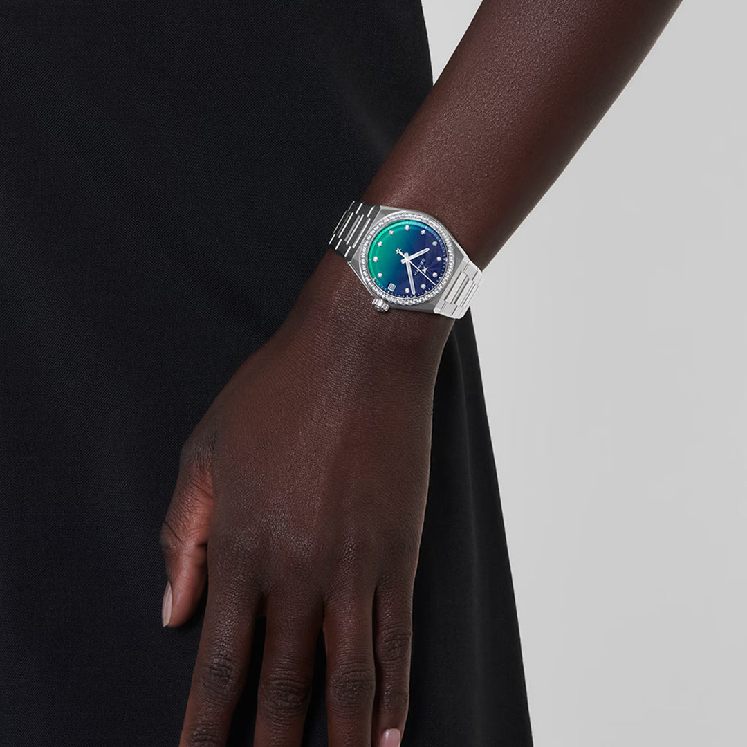 Close-up of an individual wearing a Zenith ZENITH Defy Midnight Elite 670 36mm watch, its silver bracelet complementing the green and blue gradient face. The piece features an ELITE automatic calibre, presenting a simple yet elegant design against a dark background that captures attention effortlessly.