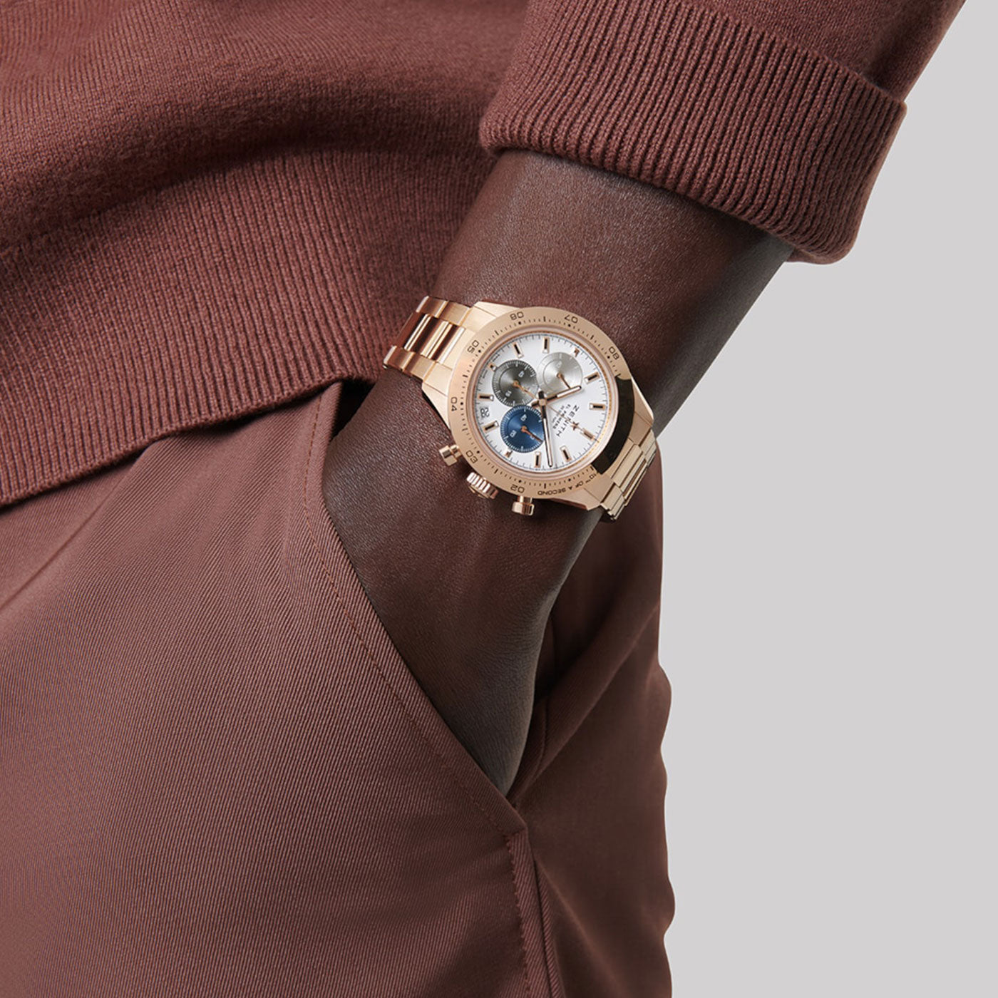 A person dressed in a brown sweater and pants has their hand in their pocket. On their wrist, the ZENITH Chronomaster Sport El Primero 3600 Automatic Chronograph 41mm Watch with a striking white and blue dial gleams elegantly against the plain gray background.