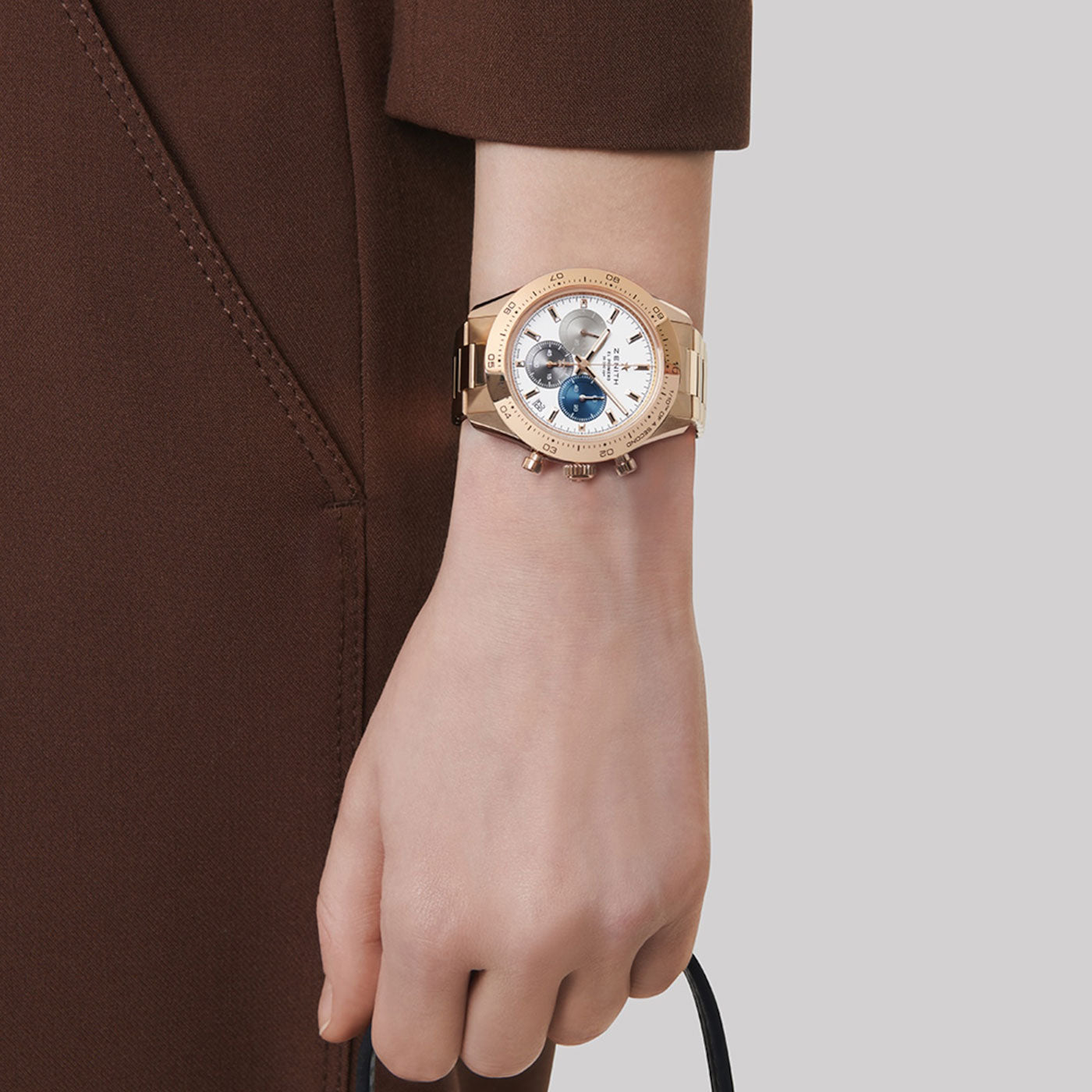 A person in a brown outfit showcases a ZENITH Chronomaster Sport El Primero 3600 Automatic Chronograph 41mm Watch, featuring multiple dials and a polished metallic band. This Zenith timepiece boasts a circular face with intricate detailing, adding elegance to the overall look against a neutral background.