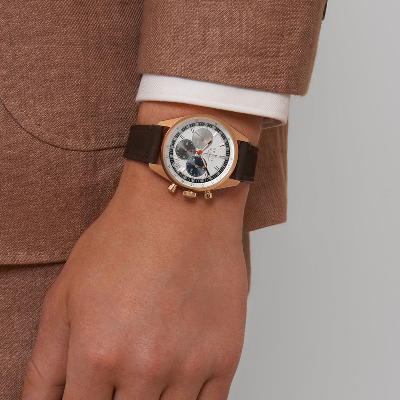A person in a brown suit and white shirt sports the chic Zenith Chronomaster Original El Primero 3600 Automatic Chronograph 38mm Watch on their wrist. This stylish timepiece features a round face accented with gold, silver, and dark blue details, all complemented by a sleek brown leather strap.